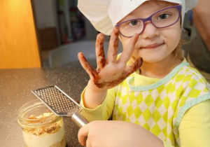 Dziewczynka z tarką w ręku podczas ozdabiania swego deseru czekoladą, której ślady widać na drugiej ręce.